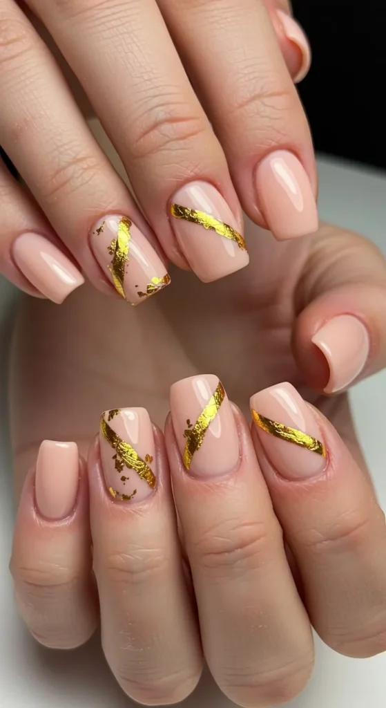 Soft peach or nude nails with metallic gold foil accents running diagonally, adding a chic and luxurious touch.

