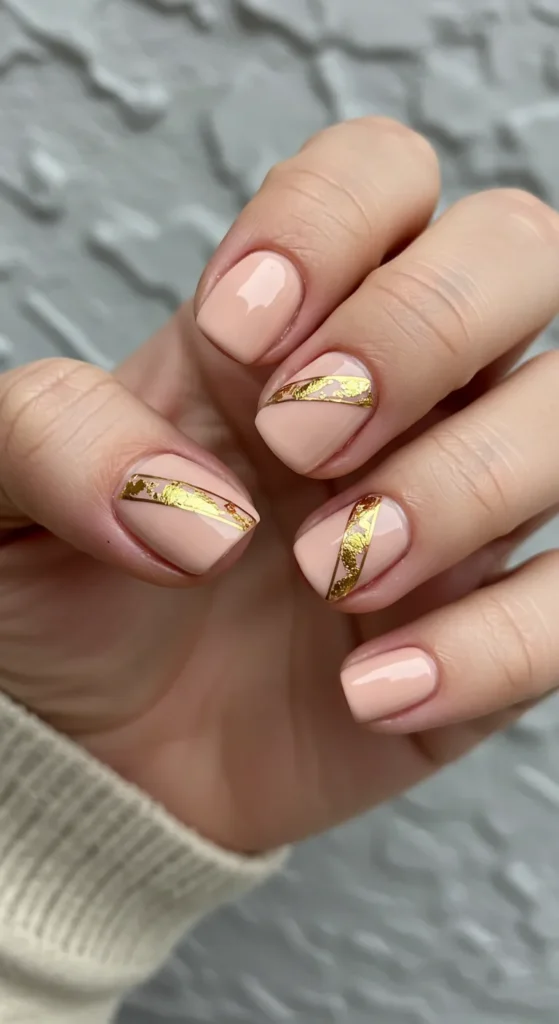 Soft peach or nude nails with metallic gold foil accents running diagonally, adding a chic and luxurious touch.

